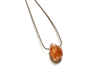 May include: A gold necklace with a teardrop-shaped sunstone pendant. The sunstone is a natural gemstone with a warm orange colour and a shimmery, iridescent finish.