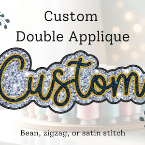 May include: Custom double appliqué with the word "Custom" in sparkly silver and navy blue lettering with a gold outline. The appliqué is on a white background with spools of thread in the foreground.