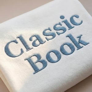 May include: A folded, off-white blanket with the words "Classic Book" embroidered in a light blue thread. The text is in a serif font and is centered on the blanket.