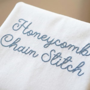 May include: Close-up of a white fabric with the words "Honeycomb Chain Stitch" embroidered in a light blue thread. The text is in a cursive font. The fabric is folded, showing a soft texture.