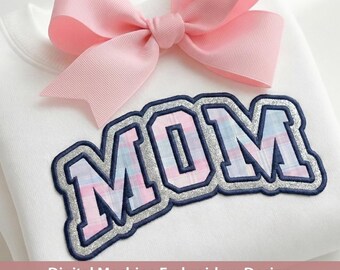 May include: A white sweatshirt featuring a large, embroidered applique of the word MOM in a plaid pattern with silver glitter borders. A large, light pink grosgrain ribbon bow is attached to the neckline of the garment.