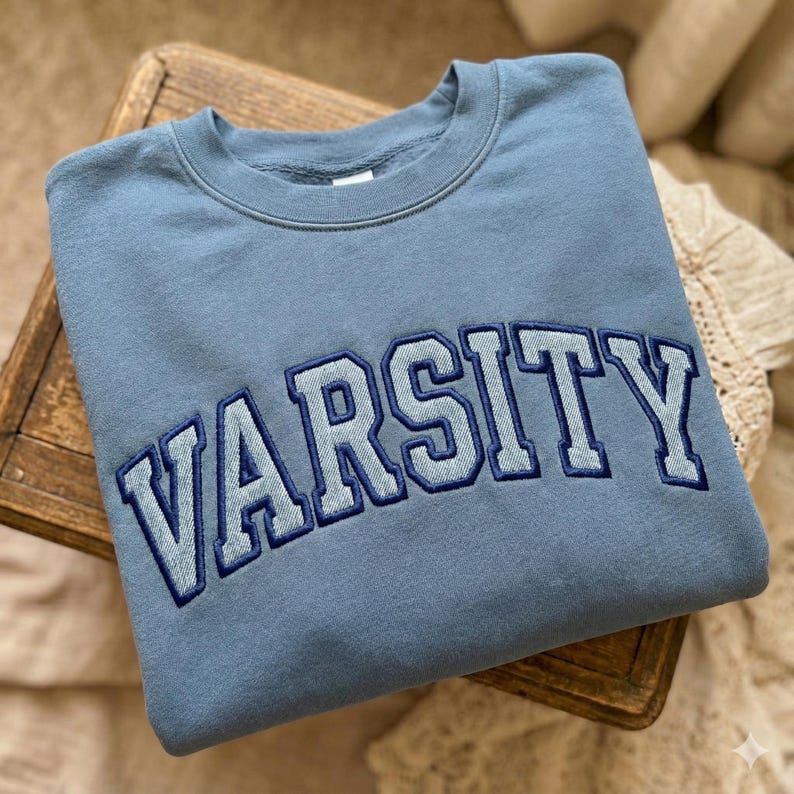 K&ouml;nnte beinhalten: Hellblaues Sweatshirt mit dem Wort "VARSITY" in dunklerem Blau auf der Brust gestickt. Das Sweatshirt liegt gefaltet auf einem kleinen Holzschemel. Der Stil ist l&auml;ssig und collegehaft.