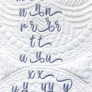 May include: A close-up of white quilted fabric with embroidered cursive letters in a soft blue hue. The letters include c, m, n, r, t, u, x, y, and z, showcasing a delicate, handcrafted aesthetic.
