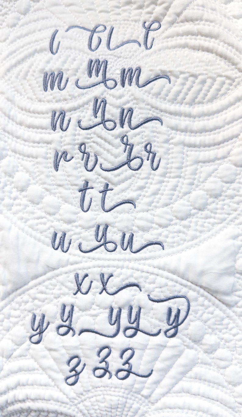 May include: A close-up of white quilted fabric with embroidered cursive letters in a soft blue hue. The letters include c, m, n, r, t, u, x, y, and z, showcasing a delicate, handcrafted aesthetic.