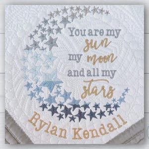 May include: A white baby blanket with a blue and gold embroidered crescent moon and stars. The text "You are my sun my moon and all my stars" is embroidered in gold. The names "Rylan Kendall" are embroidered in gold below the moon.