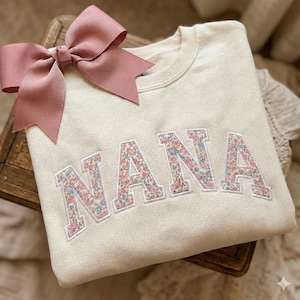K&ouml;nnte beinhalten: Cremefarbenes Sweatshirt mit dem Wort "NANA" aus floralem Stoff gestickt. Eine gro&szlig;e rosa Schleife befindet sich oben links auf dem Sweatshirt. Das Sweatshirt ist auf einer Holzoberfl&auml;che gefaltet.