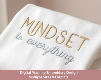 May include: A white fabric surface featuring machine embroidery with the word MINDSET in gold thread and the words is everything in light blue thread. The text is centered and stitched in a clean, sans-serif font style.