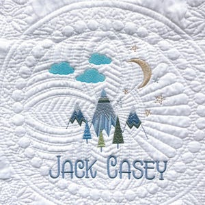 May include: A white baby quilt with a blue and green mountain scene embroidered on it. The quilt also features a crescent moon, stars, and clouds. The name "JACK CASEY" is embroidered at the bottom of the quilt.