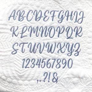 May include: A full cursive alphabet, numbers, and punctuation are shown. The soft blue letters, numbers, and symbols are embroidered onto a white, quilted background with a repeating pattern.