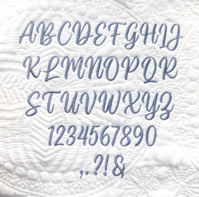 May include: A full cursive alphabet, numbers, and punctuation are shown. The soft blue letters, numbers, and symbols are embroidered onto a white, quilted background with a repeating pattern.
