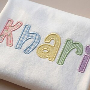Puede incluir: Tela blanca con el nombre "Khari" bordado en letras pastel. Las letras presentan diferentes patrones: floral, cuadros, lunares, rayas y un coraz&oacute;n. El bordado est&aacute; delineado en un color a juego.