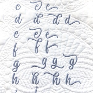 May include: A white quilted fabric featuring embroidered cursive lowercase letters in a soft grey hue. The letters a to k are shown in various styles, demonstrating different font designs. The fabric has a textured pattern.