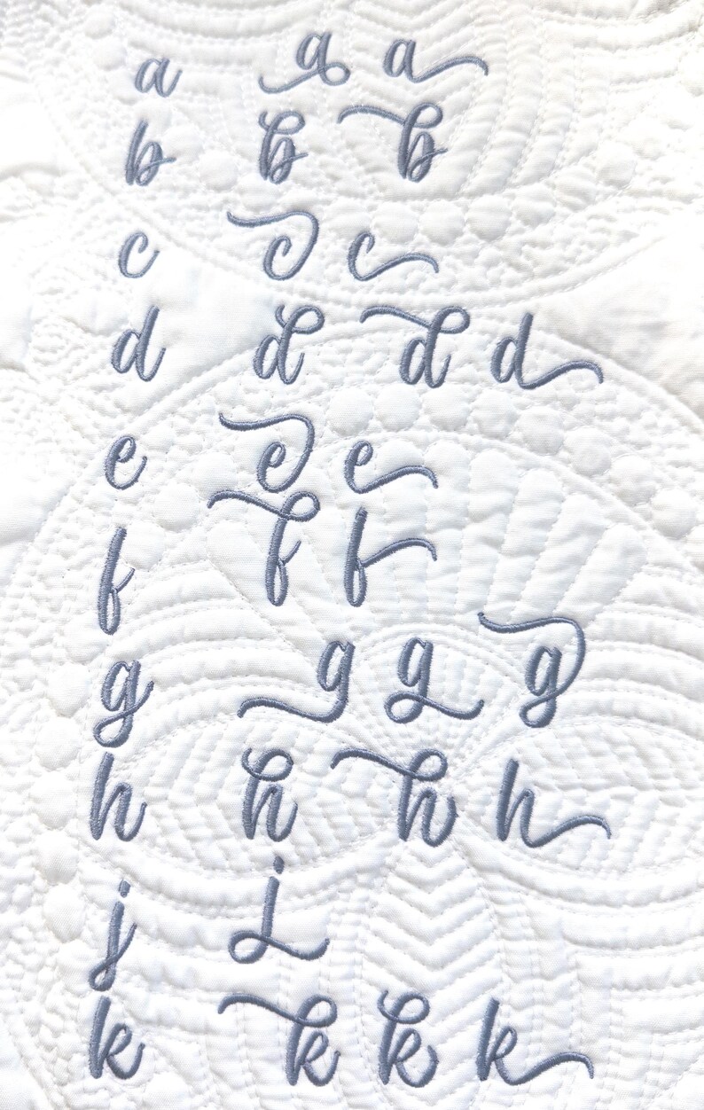 May include: A white quilted fabric featuring embroidered cursive lowercase letters in a soft grey hue. The letters a to k are shown in various styles, demonstrating different font designs. The fabric has a textured pattern.