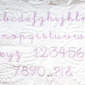 Peut inclure: Un tissu blanc avec un alphabet et des chiffres brod&eacute;s en rose. Les lettres sont en &eacute;criture cursive. Les chiffres sont dans une police similaire.