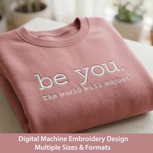 Puede incluir: Una sudadera rosa polvoriento doblada con el texto bordado "be you. The world will adjust" en blanco. La sudadera está hecha de un material suave y está doblada cuidadosamente. La imagen también incluye el texto "Digital Machine Embroidery Design Multiple Sizes & Formats."