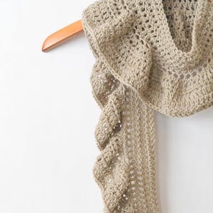May include: A beige, handmade crochet scarf with a ruffled edge, hanging on a wooden hanger. The scarf is made of a soft, textured yarn and has a delicate, open weave design. The neutral color makes it a versatile accessory.