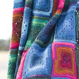 Puede incluir: Una manta de ganchillo colorida con cuadrados en tonos de azul, verde, rosa y naranja. La manta está doblada y sostenida por una persona.