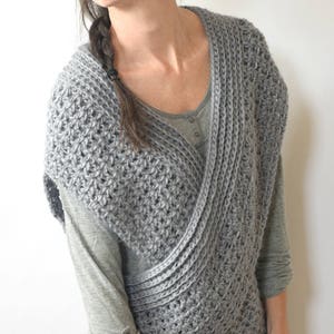 May include: A gray crocheted vest with a criss-cross design. The vest is worn over a long-sleeved shirt and jeans.
