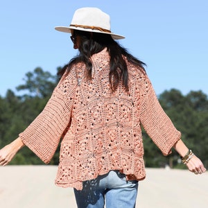 May include: A woman wearing a light pink crochet cardigan with a white crocheted hat and blue jeans.
