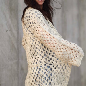 May include: A white crochet cardigan with a square pattern. The cardigan has long sleeves and a loose fit.