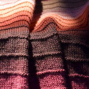 May include: Close-up of a knitted textile with a gradient of colors. The fabric transitions from light peach to dark brown, with shades of red and pink. The texture is visible, showing the individual stitches.
