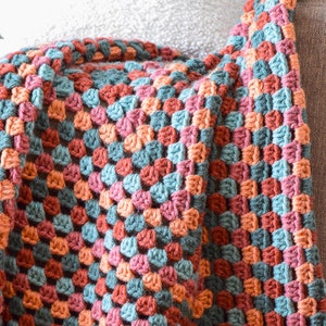 May include: A crocheted throw blanket in shades of teal, peach, and rust. The blanket is made with a granny square stitch and has a textured, woven look.