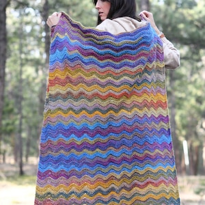 May include: A colorful crochet blanket with a wavy pattern in shades of blue, purple, yellow, and brown. The blanket is being held by a person in a forest setting.
