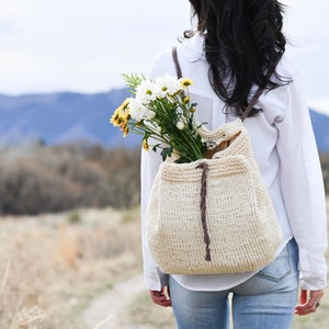 May include: A woman wearing a white shirt and blue jeans walks with a crocheted bag on her back. The bag has a brown leather strap and is filled with a bouquet of white and yellow flowers.