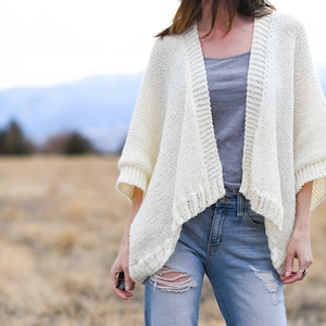 May include: A white knitted cardigan with a draped open front and 3/4 sleeves. The cardigan is worn over a gray tank top and blue jeans.