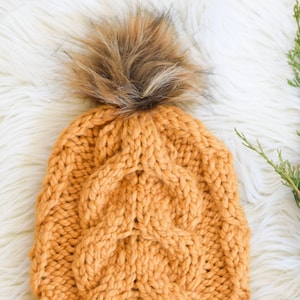 May include: A mustard yellow knitted beanie with a brown faux fur pom pom. The beanie has a cable knit pattern.