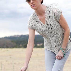 Puede incluir: Una mujer lleva una blusa de crochet beige claro con cuello en V y mangas cortas. Est&aacute; de pie en un campo con un fondo borroso de &aacute;rboles y colinas.