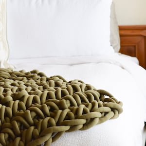 May include: A chunky knit throw blanket in a deep olive green color, draped over a white bed with a white pillow.