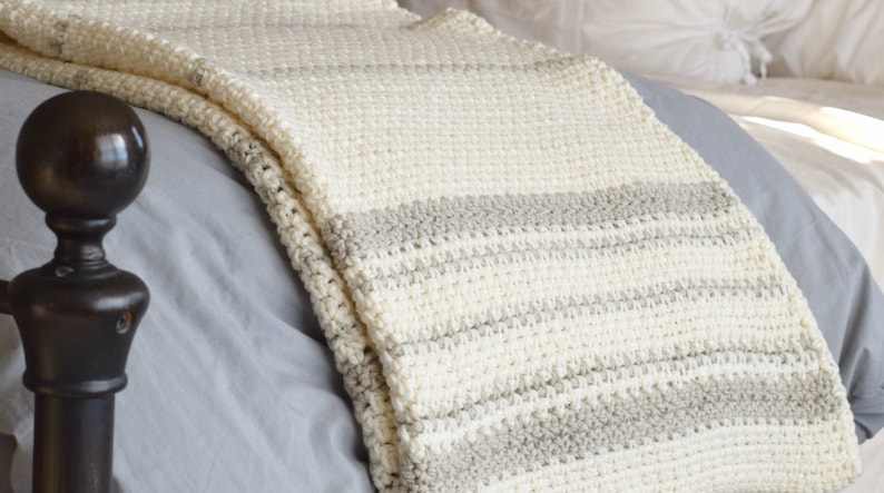 May include: A white and beige striped crocheted throw blanket draped over a bed.