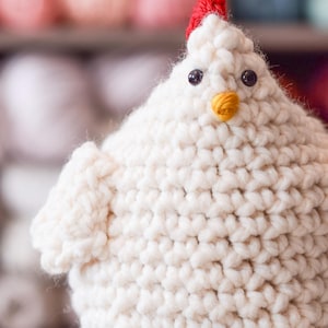 Puede incluir: Un juguete de pollo blanco de crochet con ojos negros, un pico amarillo y una cresta roja en la parte superior de la cabeza.