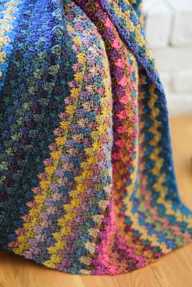 Pu&ograve; includere: Una coperta all'uncinetto multicolore con un motivo testurizzato. La coperta &egrave; piegata a met&agrave; e i colori sono blu, verde, giallo, rosa e marrone.