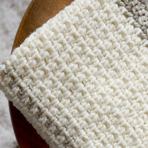May include: A close-up of a white crocheted blanket with a beige stripe. The blanket is made with a textured stitch and has a soft, cozy look.
