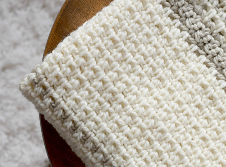 May include: A close-up of a white crocheted blanket with a beige stripe. The blanket is made with a textured stitch and has a soft, cozy look.