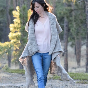 Può includere: Una donna indossa un lungo cardigan di uncinetto grigio con una camicetta rosa chiaro e jeans blu. Sta camminando in un ambiente boschivo.