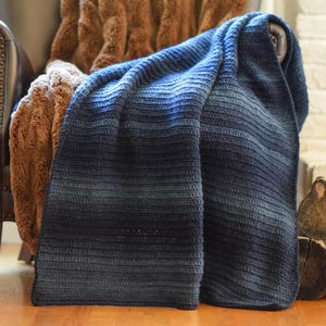 Könnte beinhalten: Eine rechteckige Häkeldecke mit einem horizontalen Farbverlauf, der von Hellblau zu tiefem Marineblau übergeht. Das Textil weist eine durchgehend gerippte Struktur auf und zeigt ein handgefertigtes Maschenbild mit weicher Optik.