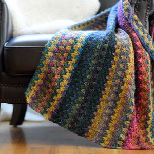 Pu&ograve; includere: Una coperta colorata all'uncinetto drappeggiata su una poltrona in pelle marrone. La coperta presenta un motivo a righe in tonalit&agrave; di blu, verde, giallo e rosa.