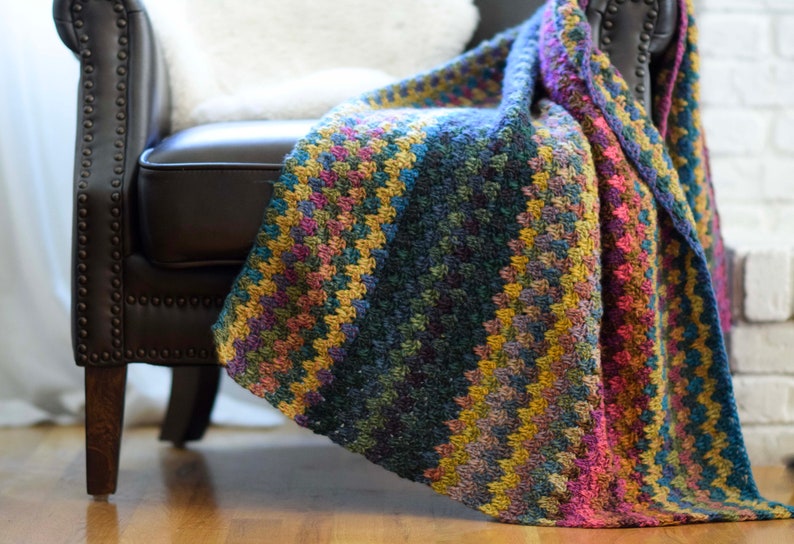 Pu&ograve; includere: Una coperta colorata all'uncinetto drappeggiata su una poltrona in pelle marrone. La coperta presenta un motivo a righe in tonalit&agrave; di blu, verde, giallo e rosa.