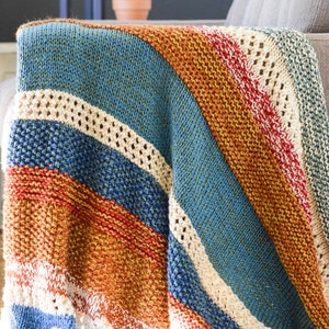 Pu&ograve; includere: Una coperta di maglia a righe in tonalit&agrave; di blu, arancione, rosso e bianco. La coperta &egrave; piegata e drappeggiata su un divano.
