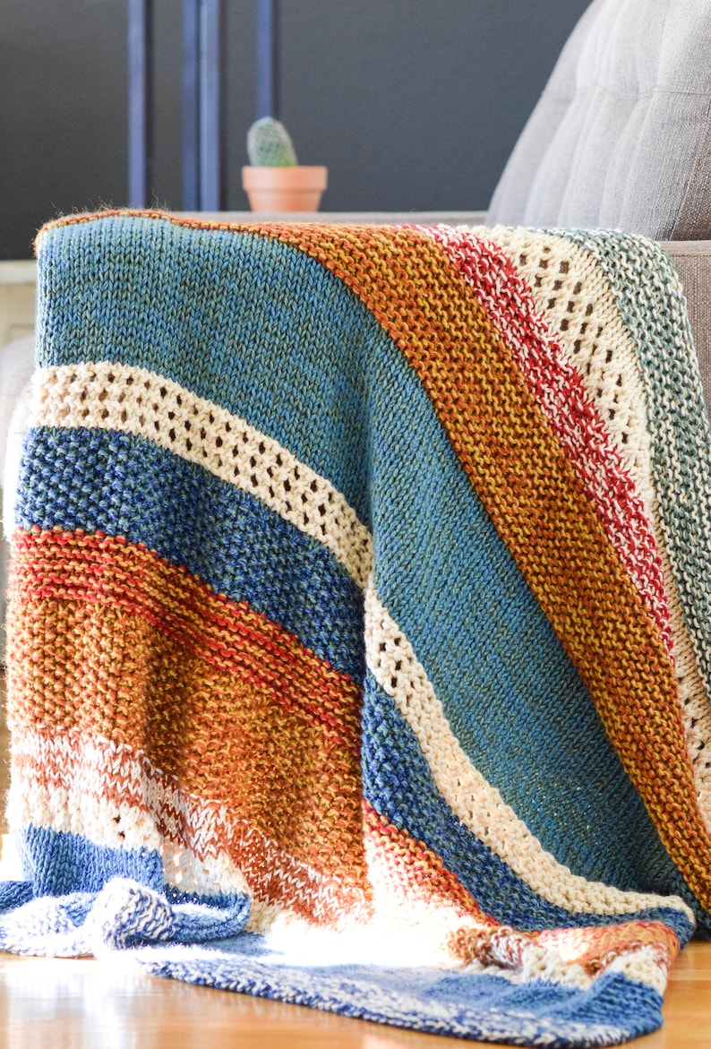 Pu&ograve; includere: Una coperta di maglia a righe in tonalit&agrave; di blu, arancione, rosso e bianco. La coperta &egrave; piegata e drappeggiata su un divano.