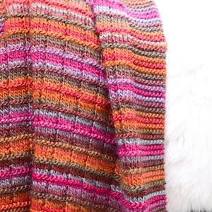 May include: A striped knitted throw blanket in shades of pink, orange, brown, and gray. The blanket is folded and draped over a white surface.