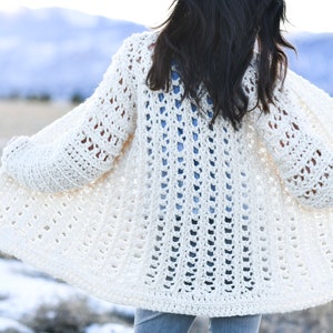 Puede incluir: Un cárdigan de crochet blanco con un patrón de punto abierto de encaje. El cárdigan se muestra desde la espalda y tiene mangas largas.