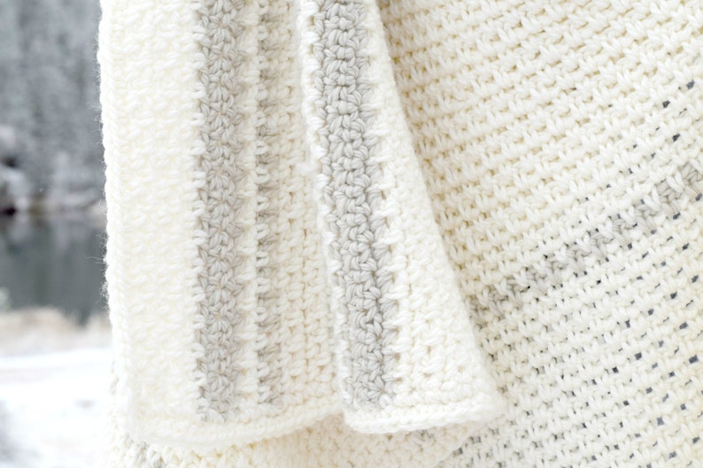 May include: A white crocheted blanket with gray stripes. The blanket is folded and draped over a surface.