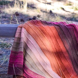 May include: A knitted blanket with a gradient of colors, from deep magenta to light pink, then to orange and brown. The blanket is draped over a wooden rail in an outdoor setting, with grass and sunlight in the background.