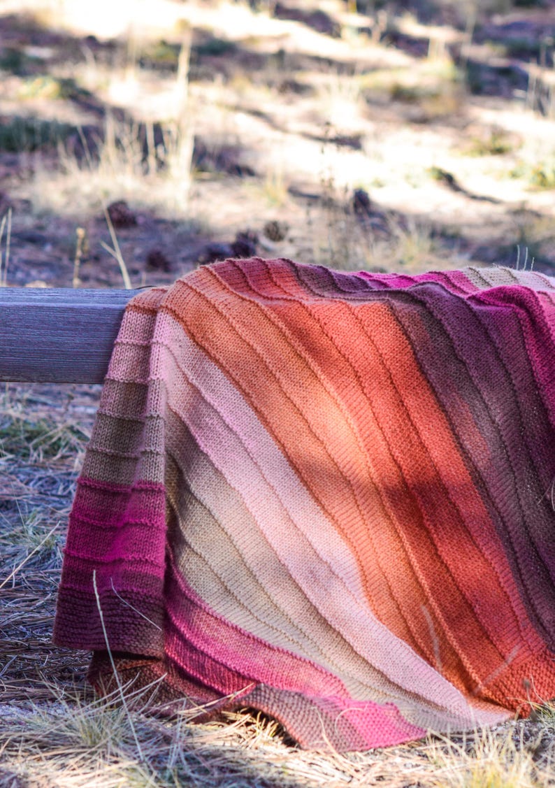 May include: A knitted blanket with a gradient of colors, from deep magenta to light pink, then to orange and brown. The blanket is draped over a wooden rail in an outdoor setting, with grass and sunlight in the background.