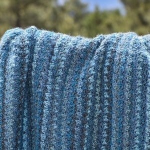 May include: Close-up of a blue and gray knitted blanket. The blanket has a textured, ribbed pattern with varying shades of blue and gray yarn. The background is blurred, showing trees and a glimpse of the sky.