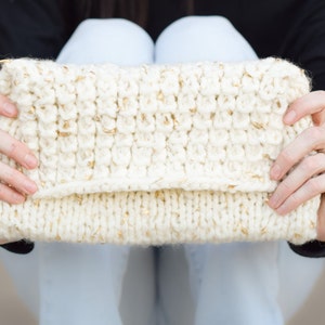 Op de afbeelding: Een witte gebreide clutch met gouden vlekken. De tas heeft een eenvoudig, gevouwen ontwerp en een gouden ringgesp.
